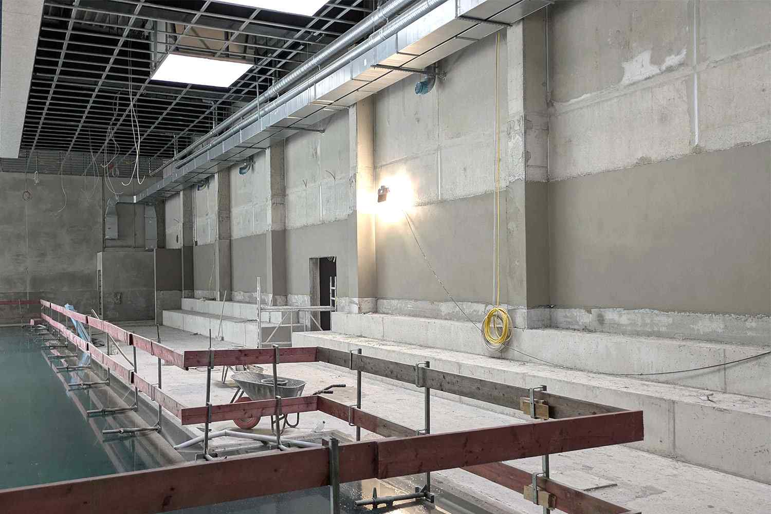 Rohbau eines Schwimmbads mit teilgefülltem Becken, Tribünenstufen und offener Decke mit sichtbaren Leitungen.